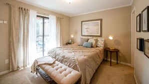 Un dormitorio con una cama grande, un banco, dos mesitas de noche con lámparas, una ventana con cortinas y cuadros en la pared.