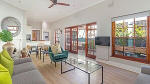 A spacious living area with a ceiling fan, a dining table with chairs, a sofa, and a large window with a view of greenery. - Gorgeous Green Point Home | 2 Bedroom | Patio (Cape Town)