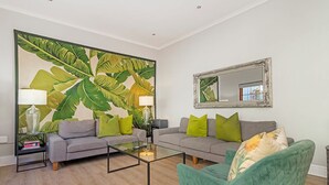 A living room with a sofa, armchair, coffee table, and a large tropical leaf painting. - Gorgeous Green Point Home | 2 Bedroom | Patio (Cape Town)