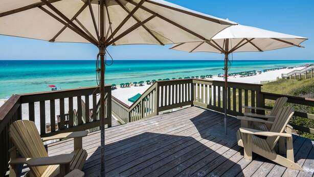 Una terraza de madera con dos sombrillas grandes, vista a una playa y una silla.