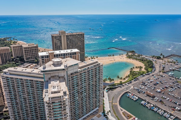 A high-rise hotel with multiple floors, a marina with boats, and a beach with palm trees.