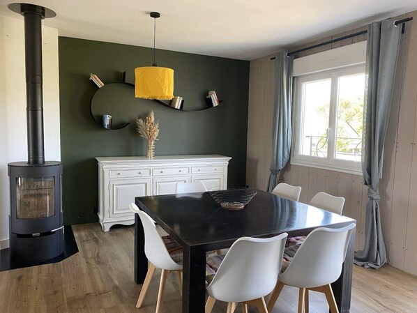 A dining area with a black table, white chairs, a white cabinet, a yellow pendant light, and a wood-burning stove.