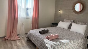 A bedroom with a bed, a bedside table, a wall-mounted mirror, and sheer curtains.