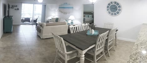 Amplia sala de estar con mesa de comedor, sillas y sofá. Se observa un reloj grande en la pared y un ventilador de techo.