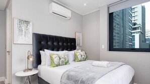 Une chambre moderne avec une tête de lit capitonnée, du linge de lit blanc et une grande fenêtre donnant sur la ville.