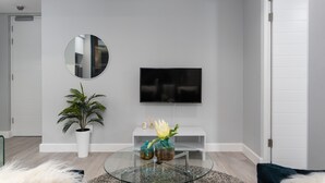 Un salon moderne avec une table basse en verre, un téléviseur à écran plat fixé au mur, une plante en pot et une console blanche.