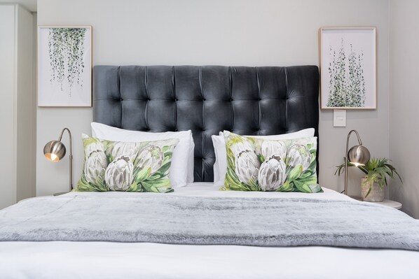 A bedroom with a tufted headboard, floral-patterned pillows, a grey shag rug, and two wall-mounted light fixtures. - Trendy Green Point Apartment with Balcony & Aircon (Cape Town)