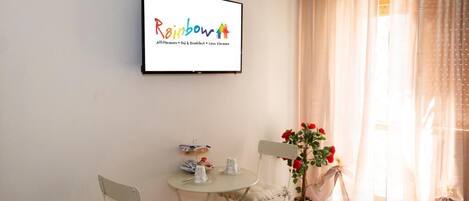 A cozy room with a framed sign reading "Rainbow," a small round table with chairs, a potted plant with red flowers, and sheer curtains.
