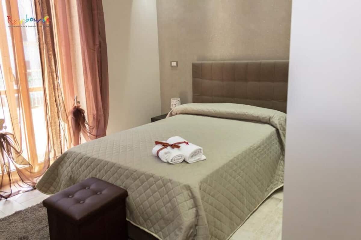 A hotel room with a bed, a nightstand, a small table, and a window with curtains.
