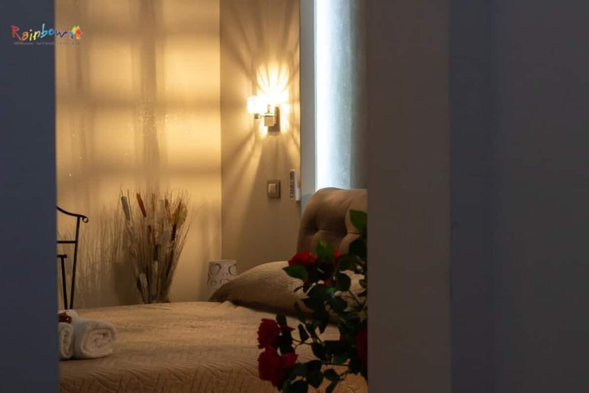 A bedroom with a bed, a bedside lamp, a vase with dried plants, and a window with sunlight.