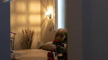 A bedroom with a bed, a bedside lamp, a vase with dried plants, and a window with sunlight.