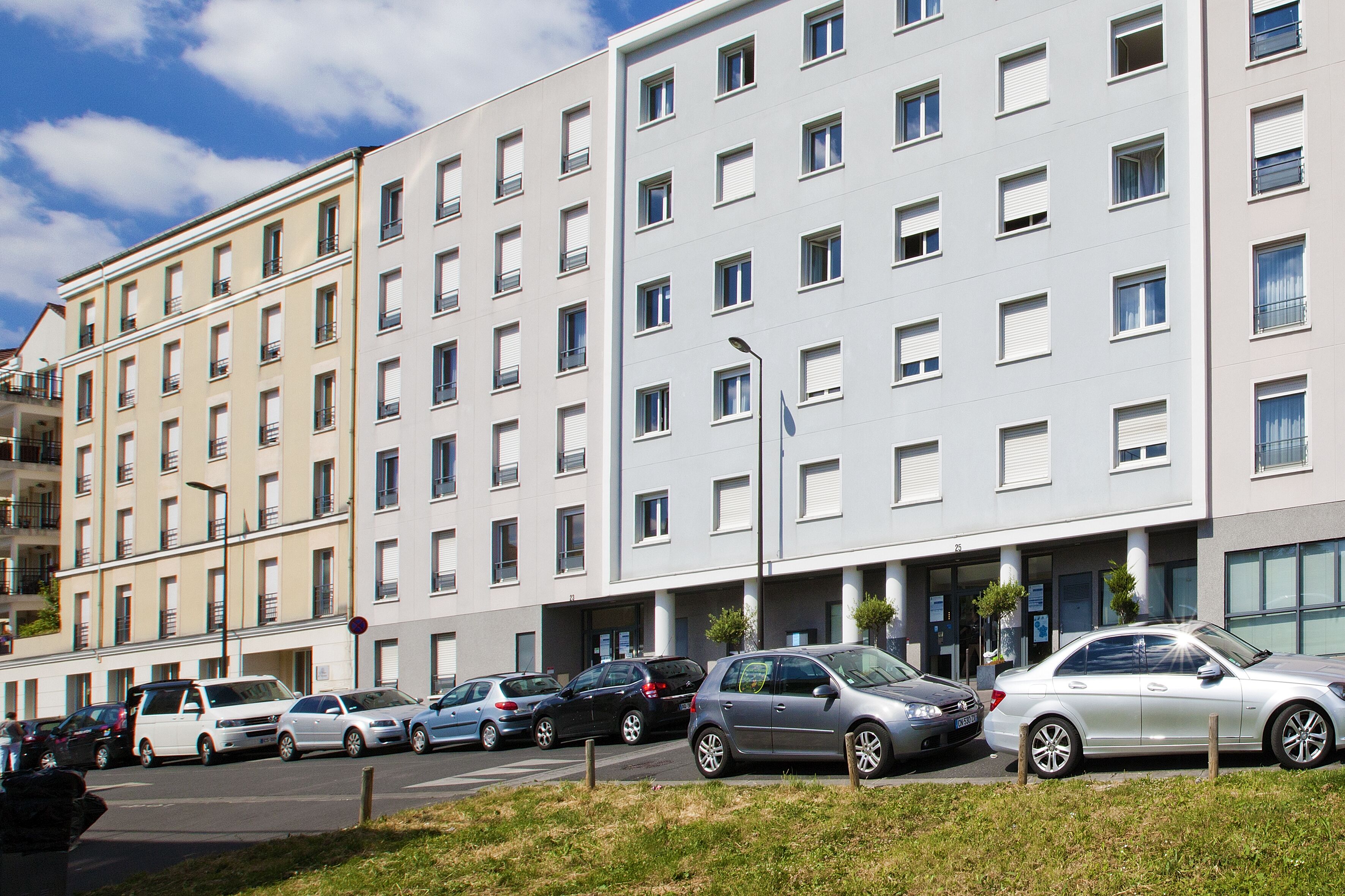Egy többemeletes lakóépület, melynek előtti járdán sorban parkolnak az autók.