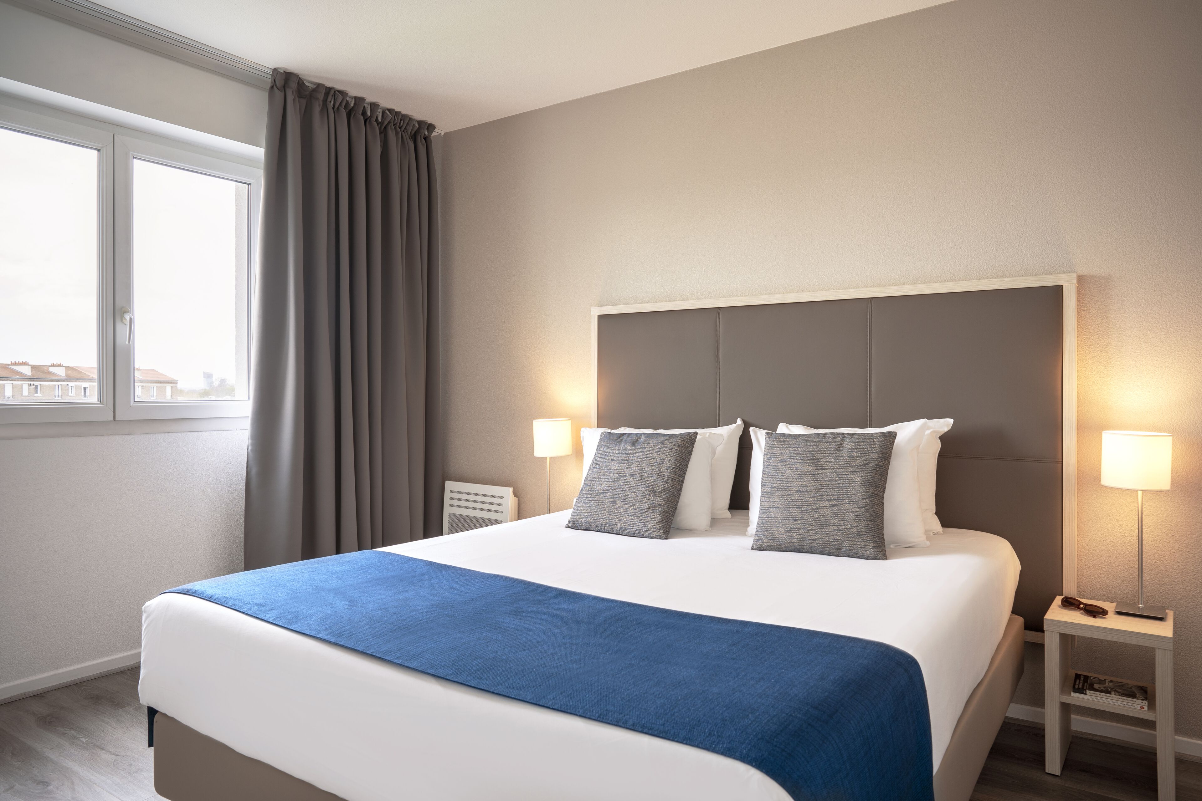 A hotel room with a large bed, two bedside lamps, a window with curtains, and a view of buildings outside.