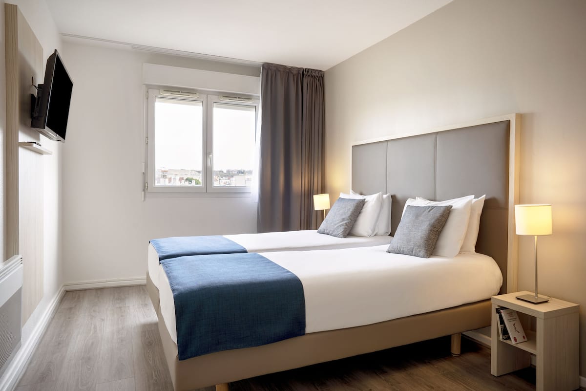 A hotel room with a large bed, two bedside tables, a TV mounted on the wall, and a window with curtains.