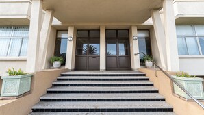 A building entrance with the name "RHODORA" above the doors. There are steps leading up to the entrance, and potted plants are placed on either side.