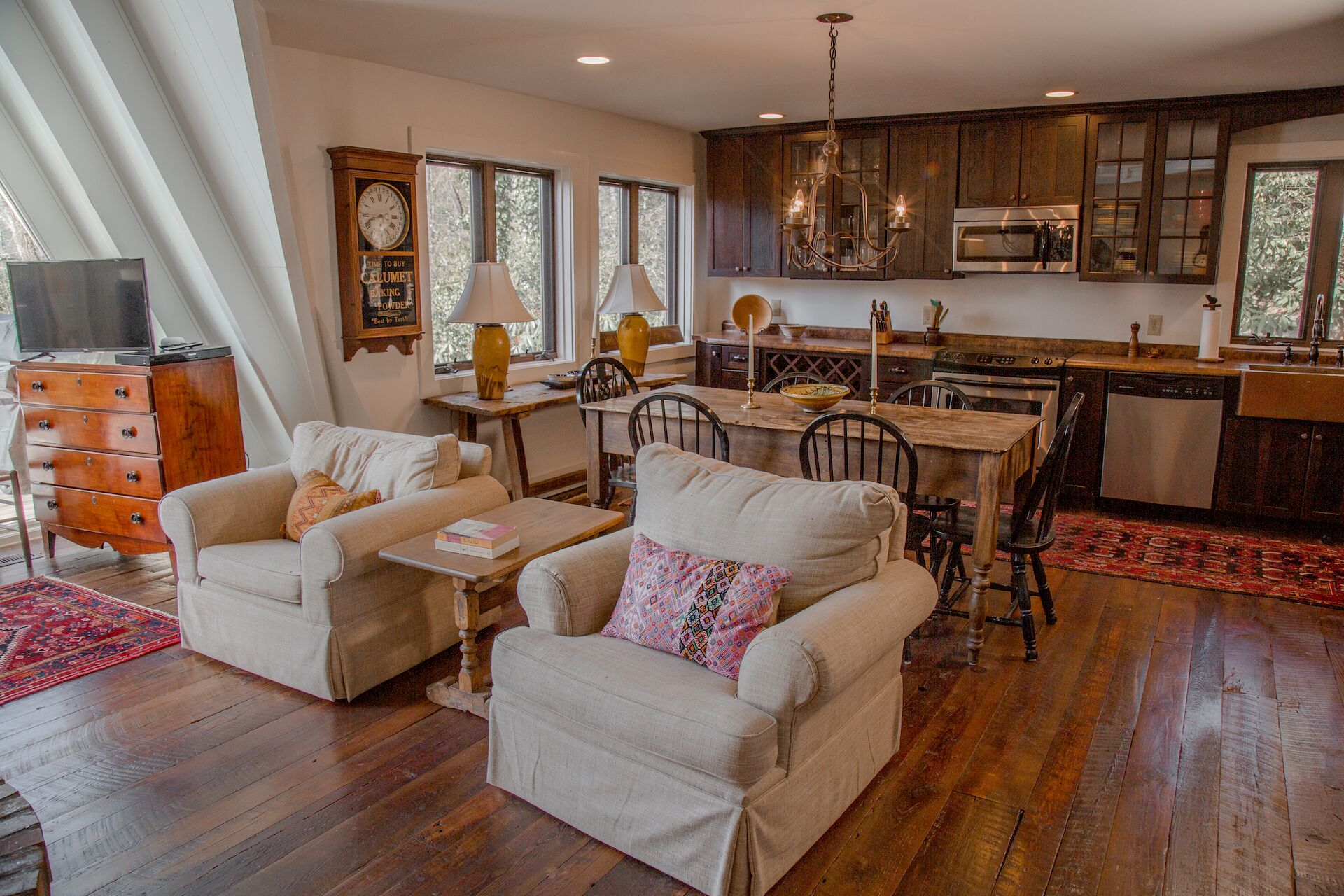 A spacious living room with a wooden floor, two armchairs, a dining table, and a kitchen area with wooden cabinets and modern appliances.