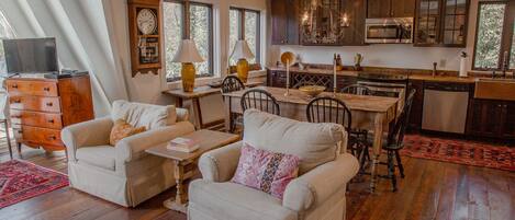 A spacious living room with a wooden floor, two armchairs, a dining table, and a kitchen area with wooden cabinets and modern appliances.