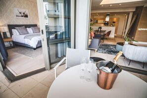 A modern hotel room with a bed, a dining area, and a kitchen in the background. There is a table with a champagne bottle and glasses. - Elegant Waterfront Apartment with No Loadshedding (Cape Town)