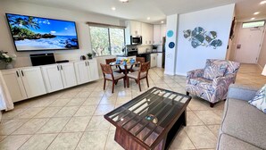 Een moderne keuken met witte kastjes, een eethoek met een glazen salontafel, en een woonkamer met een gestreept fauteuil.