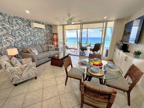 A living room with a sofa, coffee table, and dining table set with chairs. There is a balcony with a view of the ocean.