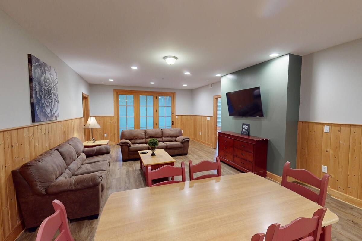 A spacious living area with a brown leather sofa, a wooden dining table with red chairs, a small side table with a lamp, and a television mounted on the wall.