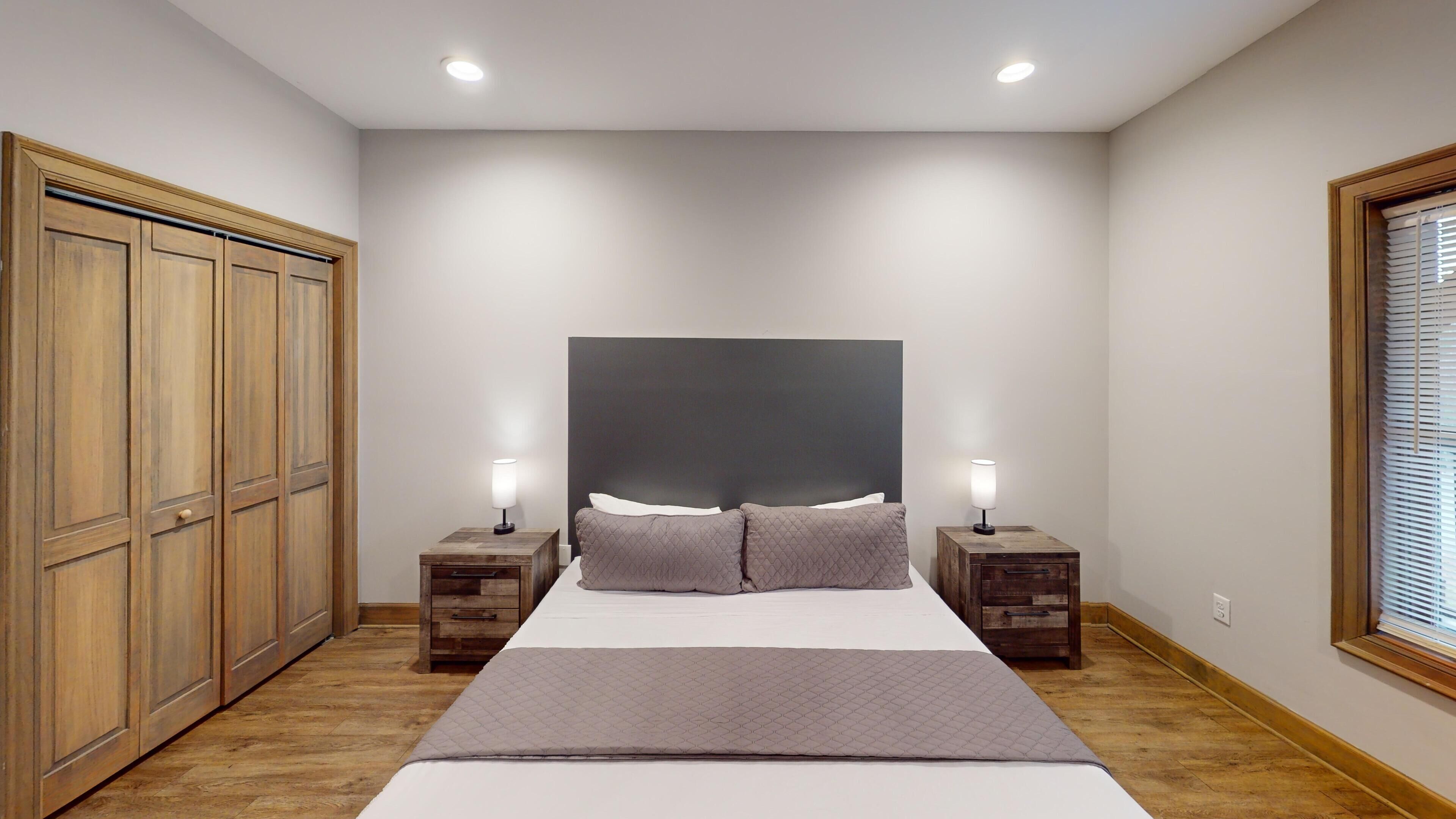A bedroom with a large bed, two bedside tables with lamps, a wardrobe, and a window with blinds.