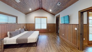 A bedroom with a wooden ceiling, a bed, a nightstand, a window with blinds, and a door leading to another room.