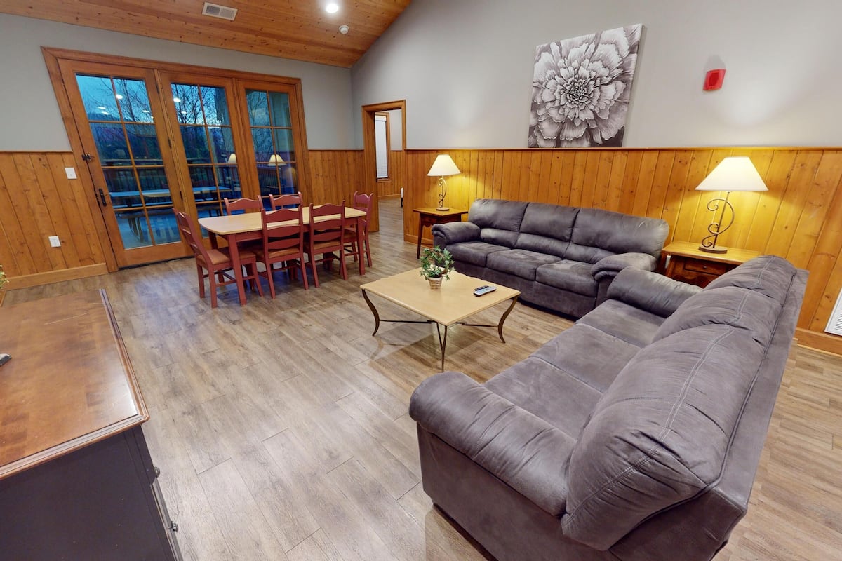 A spacious living area with a wooden floor, a grey sofa, a dining table with chairs, and a large window with a view of trees.