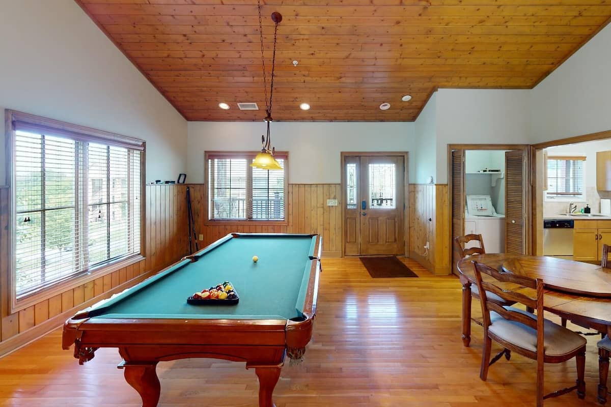 A pool table in a room with wooden floors and walls, a dining table with chairs, and a kitchen area visible through an open door.