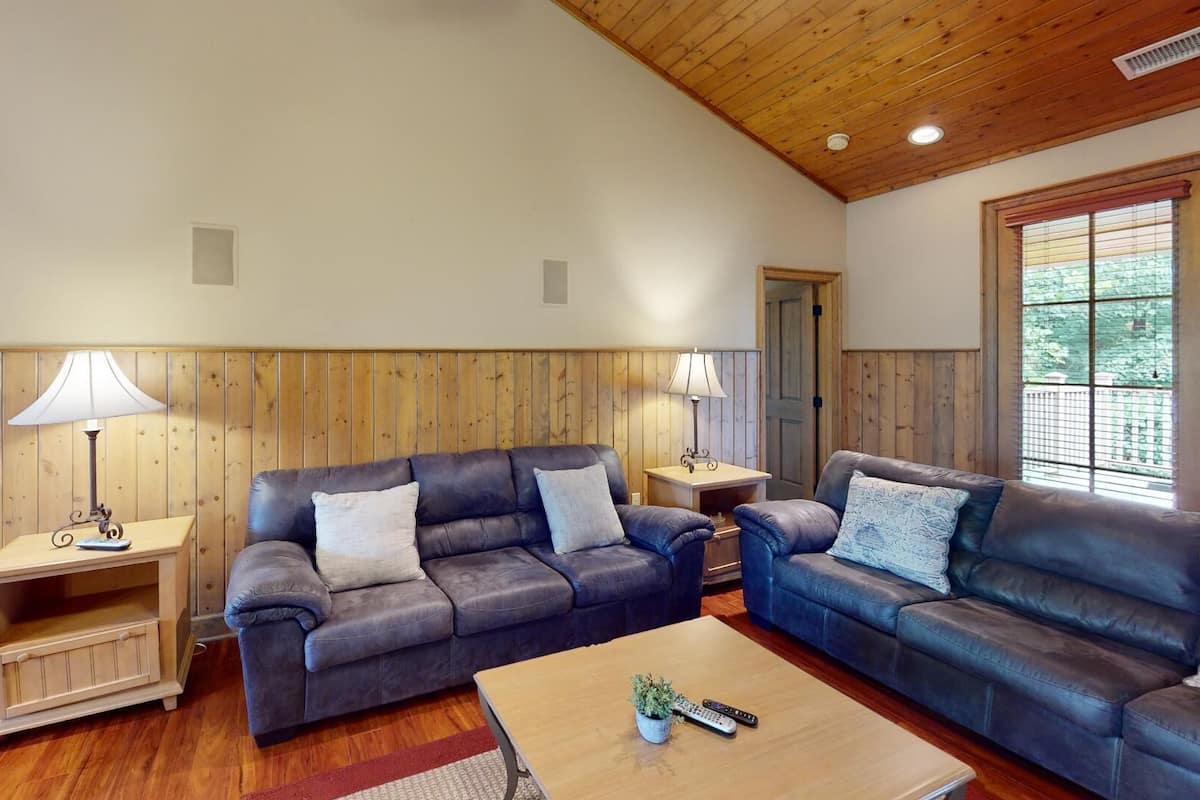 A living room with two sofas, wooden panel walls, a wooden ceiling, and a window with blinds.