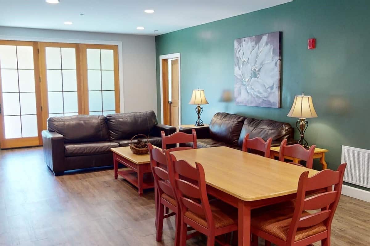 A living room with a brown leather sofa, a wooden dining table with red chairs, and a teal wall with a floral painting.