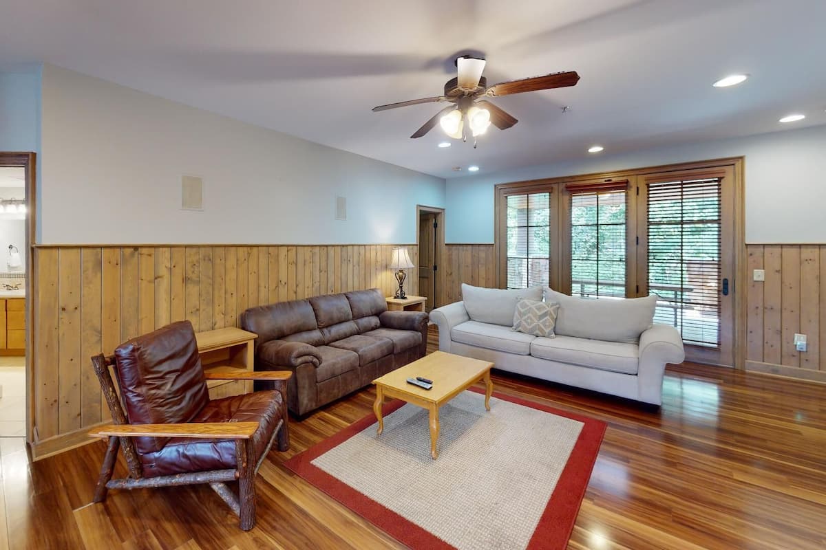 A spacious living room with a wooden floor, a brown leather sofa, a white sofa, a wooden coffee table, and a leather rocking chair.