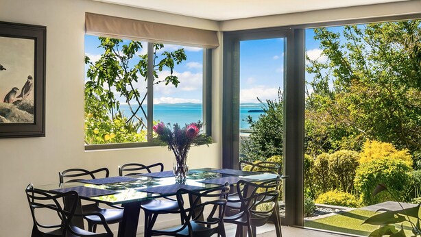 A dining area with a glass table and chairs, a vase with flowers, and a view of the sea through large windows.