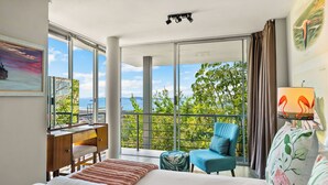 A hotel room with a large bed, a desk, a chair, and a balcony with a view of the sea and greenery.