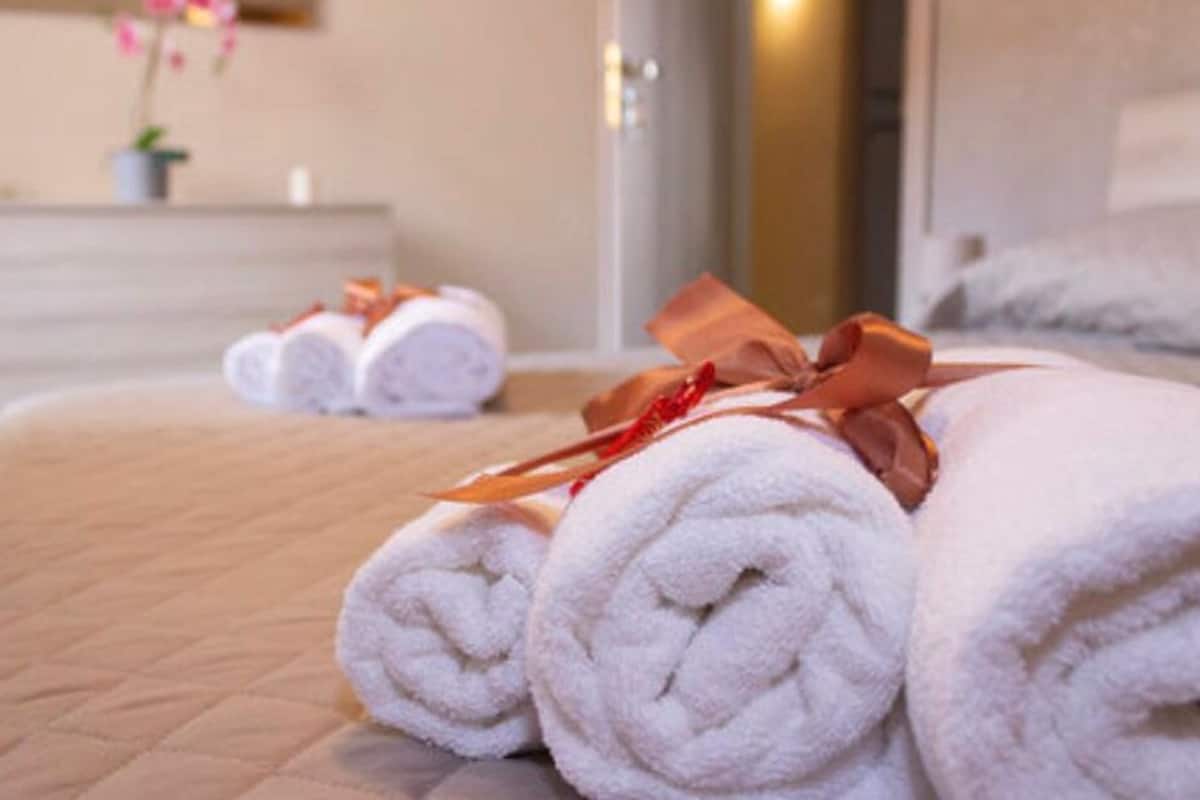 Un lit bien fait, avec des serviettes blanches roulées et un ruban rouge, une petite plante en pot et une commode en bois dans une chambre d’hôtel.