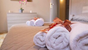 Una cama bien hecha con toallas blancas enrolladas y una cinta roja, una pequeña planta en maceta y un aparador de madera en una habitación de hotel.