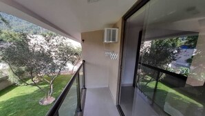 A balcony with a glass railing, a white tiled floor, and a view of a green lawn and trees. - Bugio Flats (Angra dos Reis)