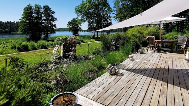 Una terraza de madera con mobiliario exterior, un dosel blanco y vistas a un lago rodeado de vegetación.