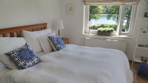 Una cama bien hecha con ropa blanca y almohadas estampadas en azul, un cabecero de madera, una lámpara de noche y una ventana con vistas a un lago.
