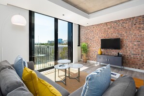 A modern living room with a grey sofa, yellow and blue cushions, a small white table, and a brick wall with a TV. - Spacious De Waterkant Apartment in The Signature (Cape Town)