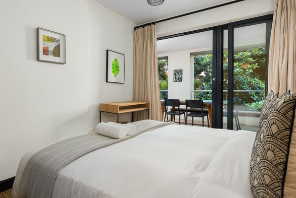 Habitación de hotel con cama, escritorio, silla y balcón con vistas a zonas verdes.