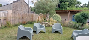 Un patio trasero con una construcción de piedra, una valla de madera y tres sillas de plástico gris.
