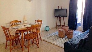 Un comedor con una mesa dispuesta para cuatro, sillas de madera, una araña, un televisor sobre un mueble bajo y un sofá al fondo.