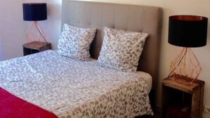 Un dormitorio con cama, dos mesitas de noche, una lámpara colgante y dos cuadros en la pared.