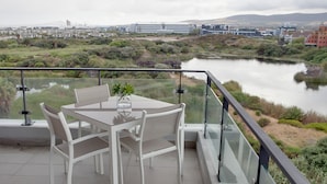 A balcony with a table and chairs, overlooking a river and cityscape. - Intaka Facing 2-Bedroom Gem (Cape Town)
