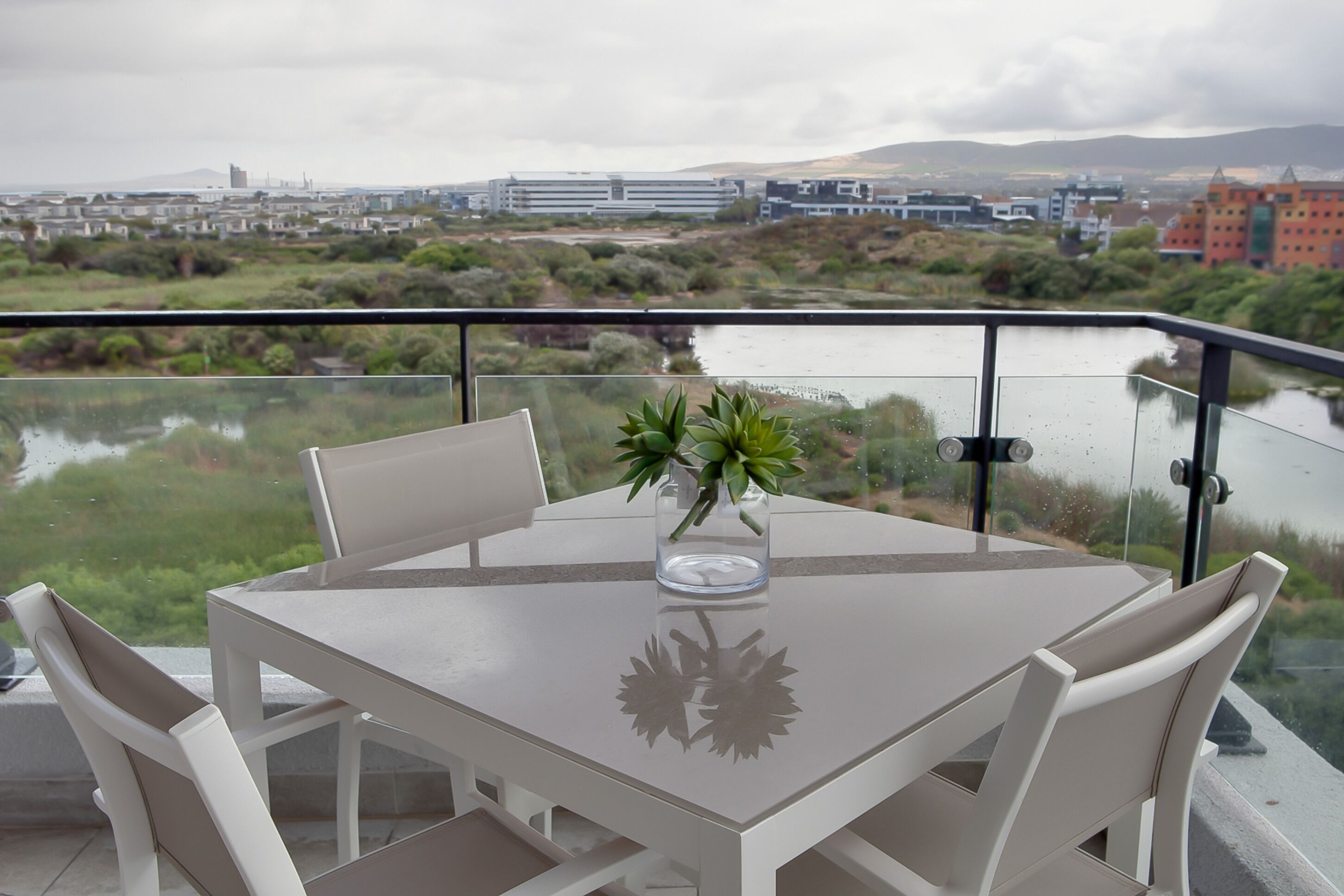 A table with chairs and a vase with plants on a balcony overlooking a body of water and buildings.