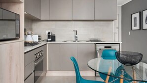 Una cocina moderna con gabinetes grises, una estufa de acero inoxidable, una mesa de comedor de vidrio y una silla azul, además de una cesta de malla negra.