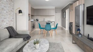 Una sala de estar moderna con un sofá gris, una mesa redonda de centro con una planta, y un comedor con sillas color turquesa y una mesa blanca.