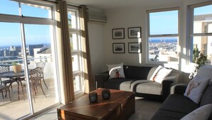 A modern living room with a sofa, coffee table, and dining area with chairs. Large windows offer a city view. - Exclusive Townhouse with Stunning Views (Cape Town)