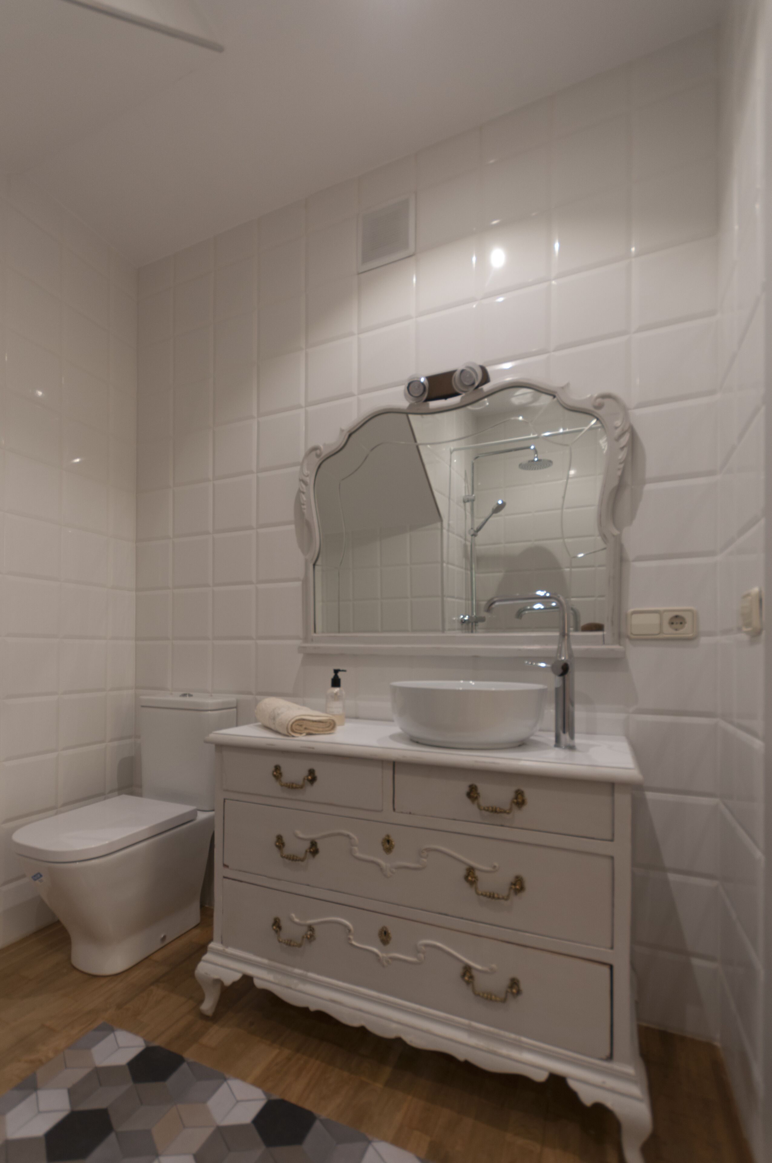 Un baño con azulejos blancos, un lavamanos blanco con un espejo encima, un inodoro y un armario blanco con cajones.